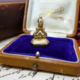 Gold decorative item on a purple cushion inside an open jewelry box with visible branding.