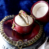 Gold ring with a red gemstone in an open wooden box on a decorative stand.