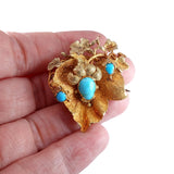 Gold brooch with turquoise stones held between fingers on a white background
