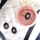 Black star-shaped earrings with gold accents on a white surface with a pink jewelry box.