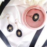 Pair of black earrings with gold accents on a white surface with a pink jewelry box.