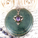 Vintage-style purple gemstone pendant on a green stone surface with white roses and lavender flowers.