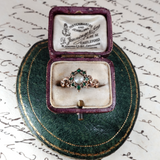 Vintage ring with emeralds and pearl in an open jewelry box on a textured surface.