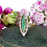 Gold ring with emerald and diamond design on a green surface with flowers in the background