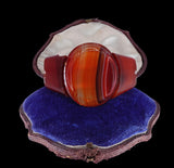 A large oval ring with a polished banded agate or jasper face in orange, red, and brown tones, displayed in an open vintage shell-shaped box lined with white material and resting on a blue velvet cushion against a black background.