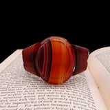 A carved wooden bowl with a rich reddish-brown and orange striped pattern, resting on the pages of an open book against a black background.