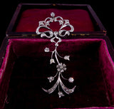 A silver-toned floral and scroll brooch embellished with small clear stones, displayed inside an open dark velvet jewelry box with a burgundy interior.