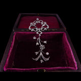 An ornate silver-toned floral vine pendant with clear stones, displayed inside an open dark brown presentation box lined with deep magenta velvet against a black background.