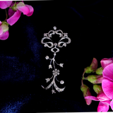A silver-toned filigree brooch with small clear stones, displayed on a black velvet background with pink flowers in the corners.