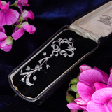 A silver-toned filigree brooch with clear stones, displayed inside an open vintage black velvet jewelry box on a dark blue velvet background, framed by pink flowers.