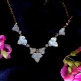 Gold-tone necklace with carved white mother-of-pearl butterfly motifs and small clear faceted accents, displayed on a dark velvet background with pink flowers at the corners.