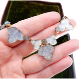 A close-up of a hand holding a vintage gold-tone necklace with carved white mother-of-pearl leaf pendants and small clear stones, with a dark green velvet jewelry box in the background.