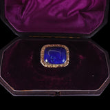 A square vintage brooch with a deep blue cabochon stone and a gold-toned beaded border, displayed inside an open dark purple velvet jewelry box on a black background.