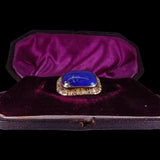 A vintage-style ring with a large blue cabochon stone in a gold-toned setting, displayed inside an open dark box with a purple velvet interior.
