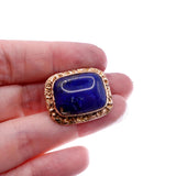 A rectangular cabochon lapis lazuli gemstone with a gold-toned filigree border, held on a person's fingers against a white background.