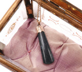 A black cylindrical pendant necklace with a rose gold cap and chain, resting on a dusty purple fabric inside a wooden box.