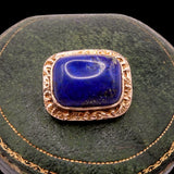 A vintage rectangular lapel pin featuring a deep blue lapis lazuli stone set in an ornate rose gold filigree frame, displayed on a dark green velvet jewelry cushion.