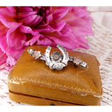 A silver-toned horseshoe-shaped hair claw clip with floral details, displayed on top of a vintage brown leather case with a large pink flower in the background.