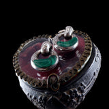 A pair of silver-toned hoop earrings with curved green stone inlays, displayed on top of an ornate heart-shaped dark metal box against a black background.