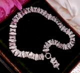 A silver-toned beaded bracelet with irregular, polished stone-like beads, displayed in a dark brown jewelry box with a black velvet lining. Pink fabric and a white satin cloth are visible in the corners.
