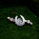 A silver-toned metal hair claw clip decorated with a central daisy motif, a curved crescent shape, and two small flower accents, displayed on a textured dark green surface.