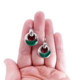 A pair of silver-toned crescent-shaped dangle earrings with green stone inlays, held between a person's fingers against a plain white background.