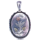 An oval pendant featuring a silver-toned metal frame with a floral engraving in the center, accented with pink and gold-colored details, set against a plain white background.