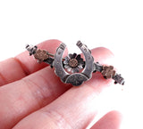 A close-up of a hand holding a small silver-toned Japanese-style brooch featuring a central flower motif, floral accents, and engraved details, set against a plain white background.