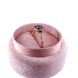 A gold ring with a round purple gemstone is displayed inside a round pink velvet ring box with a ribbed fabric lid, set against a plain white background.