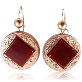 A pair of rose gold drop earrings featuring a square dark red gemstone in the center of an ornate circular rose gold frame, suspended from fishhook wires on a white background with a soft reflection.